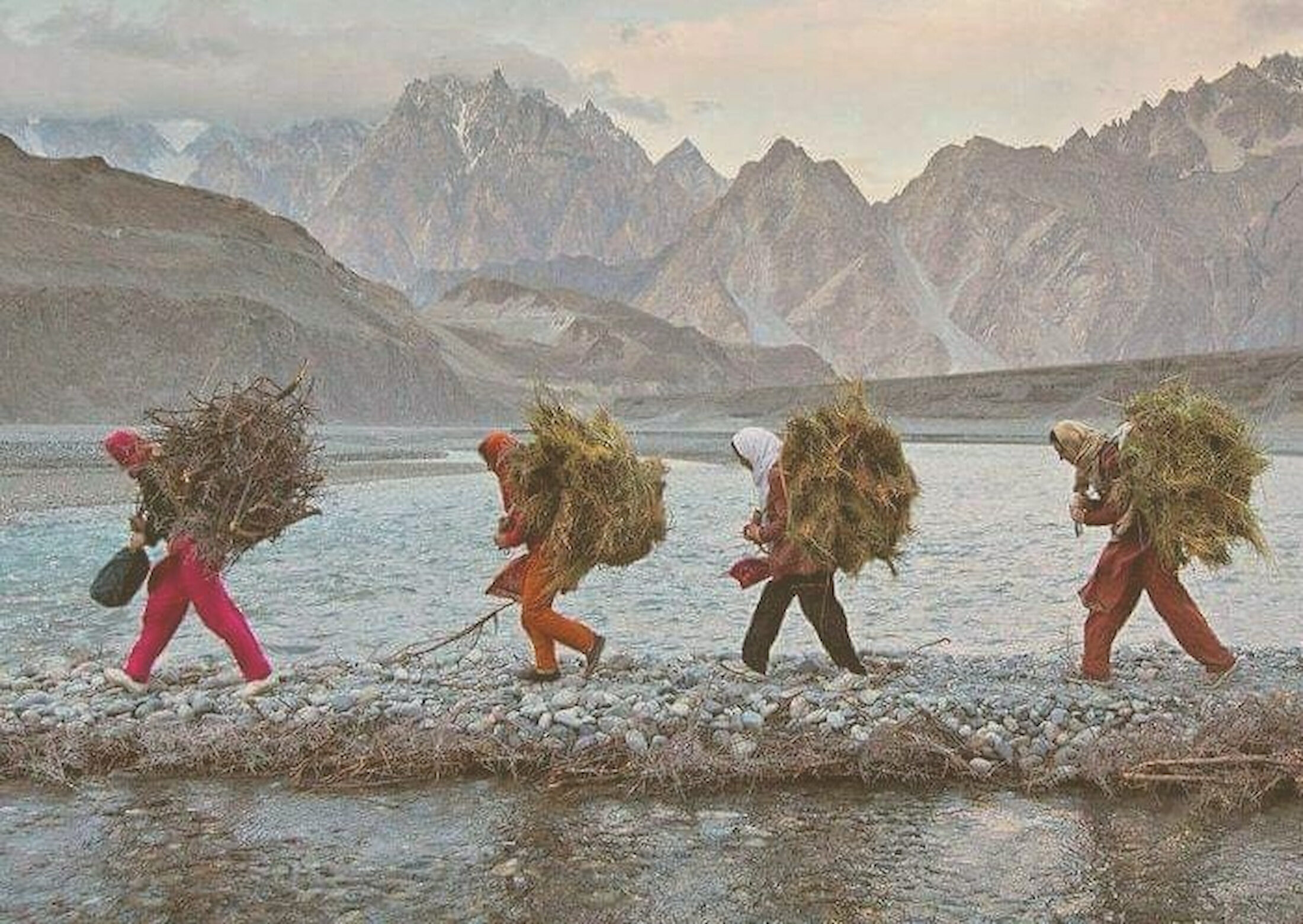 Women carrying bundles of hay along a river beneath the jagged peaks of the Karakoram